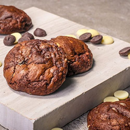 Two dark chocolate cookies with cracked tops on a pale stone slab, surrounded by dark and white chocolate buttons