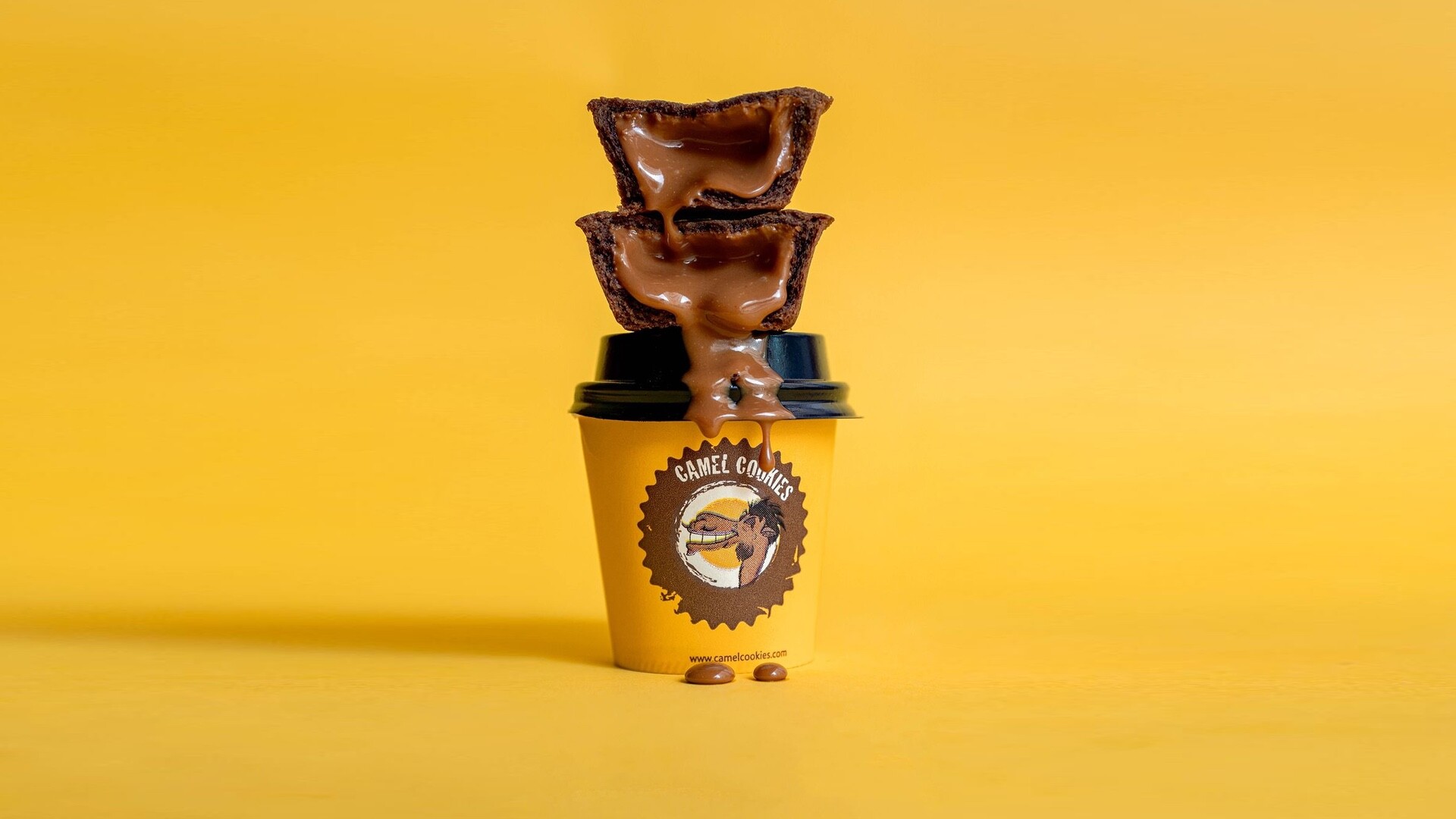 Two chocolate cookie cups stacked on a branded yellow Camel Cookies coffee cup, with molten chocolate dripping down the side