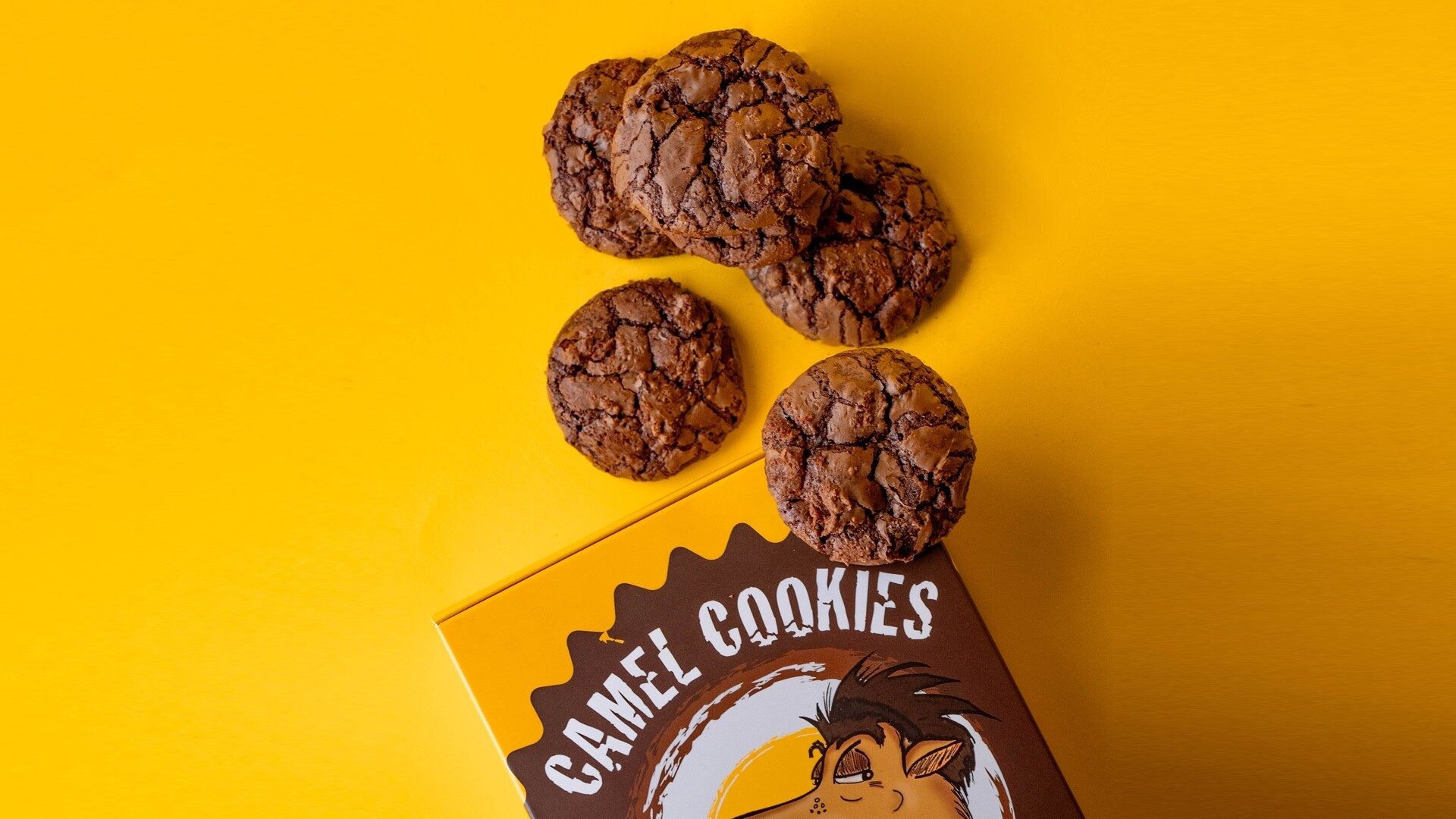Cracked-top chocolate cookies scattered beside an open yellow Camel Cookies box on a saffron-yellow surface
