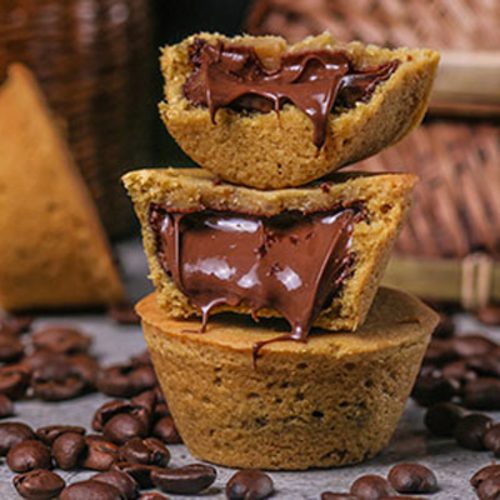 Three Camel Cookies cookie cups stacked on a stone surface scattered with roasted coffee beans, the top two split to show a glossy chocolate hazelnut centre